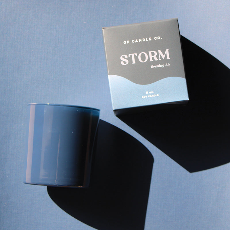 GP Candle Co. 9 oz 'Storm' candle in deep grey colored glass vessel and packaging displaying scent note "Evening Air" on a blue-grey background.