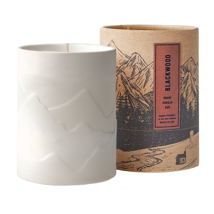 White, textured 12 oz candle vessel next to a cardboard tube packaging with mountain design and 'Chalet' text and scent notes "Ebony+Vanilla+Oud".