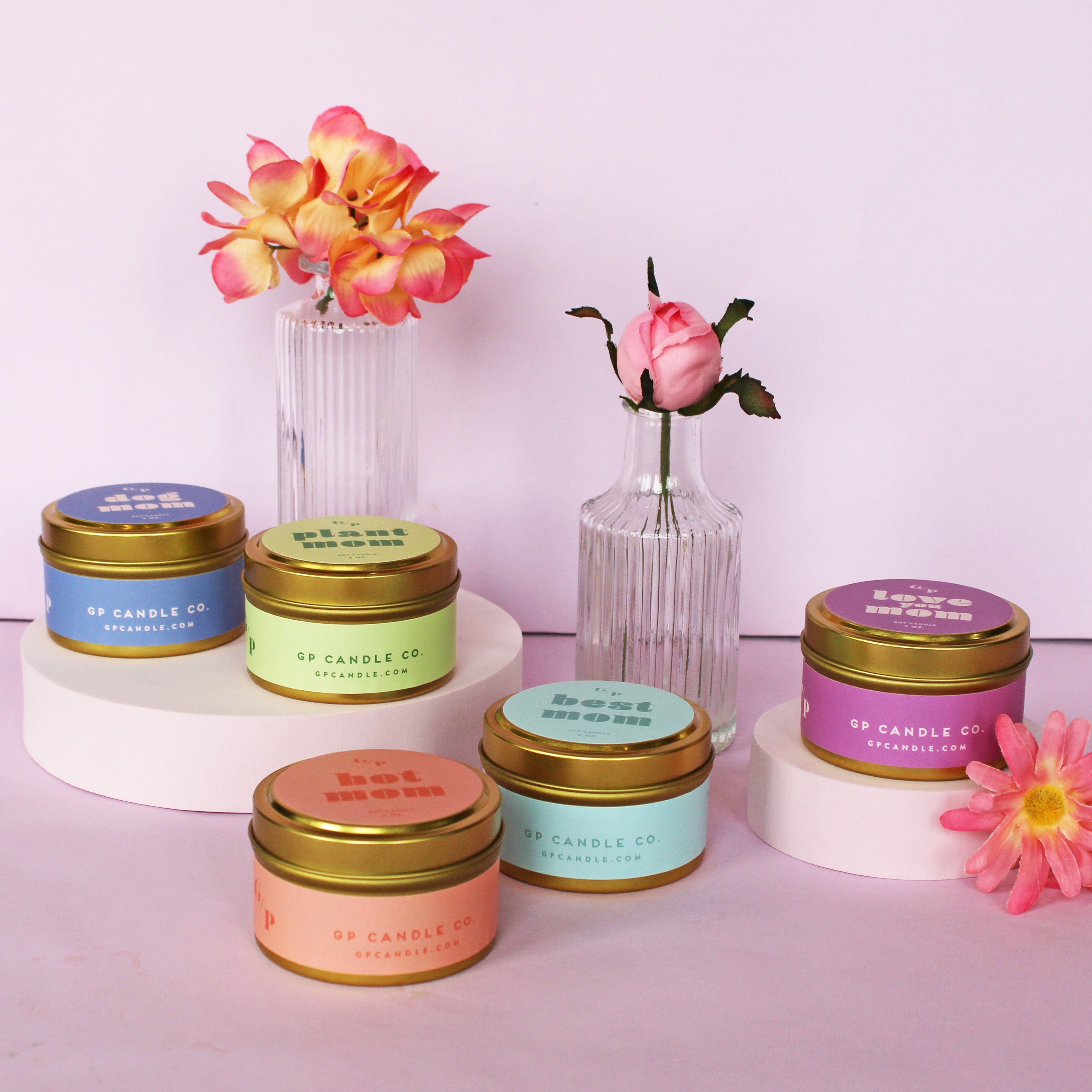 5, 4 oz candle tins labeled 'love you mom', 'dog mom', 'hot mom', 'plant mom', 'best mom',  with colorful labels on a pink background and vases holding flowers.