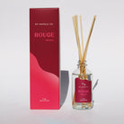 Glass diffuser bottle with wooden reeds and red box labeled 'Rouge' with the scent note of "Elderberry" displayed on a sunny white background.